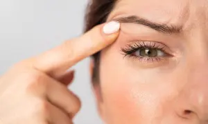 Dettaglio di un occhio incappucciato con trucco, evidenziato da un dito che solleva la palpebra.