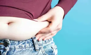 Mano che stringe la pancia, evidenziando gonfiore e possibile ritenzione idrica pancia. Jeans a vita bassa e maglia bordeaux.