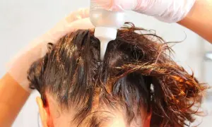 Mani guantate applicano un prodotto per capelli, forse una tinta o un trattamento, direttamente sul cuoio capelluto. Si intravede il flacone con beccuccio.