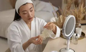 Donna con fascia in testa e accappatoio bianco tiene in mano una maschera in tessuto. Sul tavolo, uno specchio e prodotti per la cura della pelle, ideali come tonico prima o dopo la maschera.