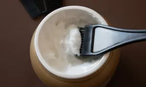 Un pennello raccoglie una crema bianca da un barattolo dorato, ideale per la cura dei capelli con burro di karitè.