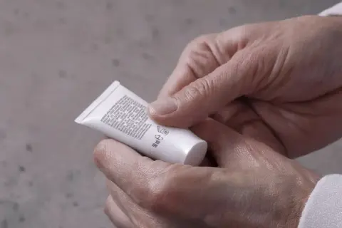 Mani che stringono un tubetto di crema, forse un trattamento iodase anticellulite tiroide, leggendo attentamente gli ingredienti.