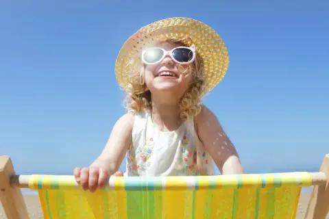 Bambina sorridente con cappello di paglia e occhiali da sole, seduta su una sdraio. Posso usare la crema solare per bambini per proteggerla dal sole.