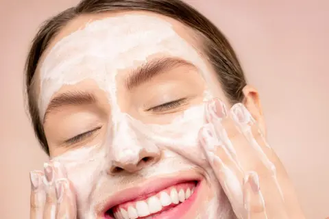 Donna sorridente con maschera bianca sul viso, mentre impara come fare una pulizia del viso a casa.
