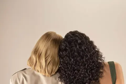 Due donne, una con capelli biondi lisci e l'altra con capelli scuri ricci, si abbracciano. La domanda è quanto dura una tinta permanente.
