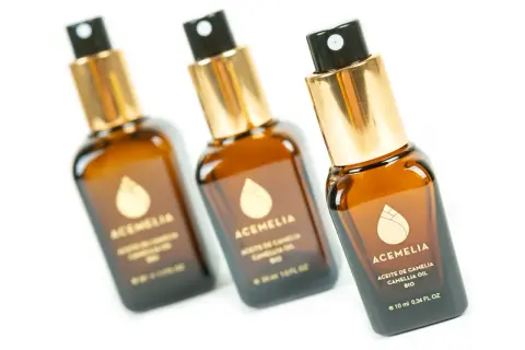Tre flaconi di olio di camelia per capelli, con tappo dorato e spruzzino nero, su sfondo bianco.