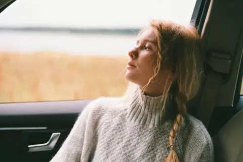 Ragazza con capelli biondi raccolti in acconciature trecce laterali, guarda fuori dal finestrino dell'auto, immersa nei suoi pensieri.