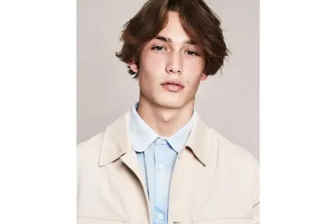 Ragazzo con capelli castani mossi e acconciature alla moda, indossa una camicia azzurra e una giacca beige.