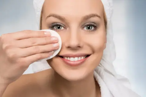 Donna sorridente con asciugamano in testa, pulisce il viso con un dischetto. Scopri i rimedi della nonna per pori dilatati.