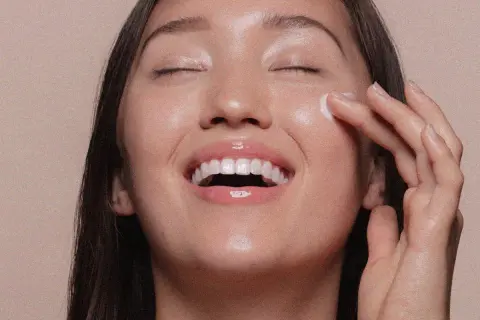 Viso radioso di una donna che applica una crema, un gesto che ricorda cos'è lo scrub per una pelle luminosa e liscia.