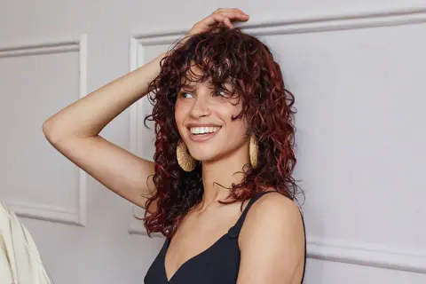 Donna sorridente con capelli castani mossi e tinta rossa, indossa orecchini dorati.