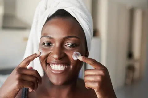 Donna sorridente con turbante applica crema sul viso, un'immagine che evoca il desiderio di vedere i risultati del **retinolo prima e dopo**.