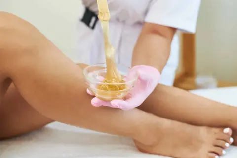 Una mano con guanto rosa preleva cera da una ciotola per la depilazione femminile delle gambe.