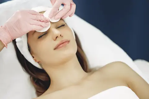Mani con guanti rosa eseguono una delicata pulizia del viso con dischetti di cotone su una donna rilassata.