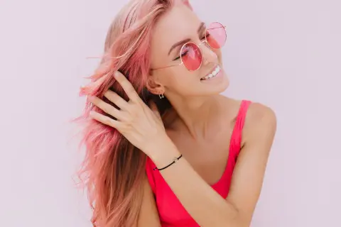 Ragazza con capelli rosa sfumati, occhiali da sole rotondi e un top fucsia. Il suo shampoo colorante mantiene vibrante la chioma.