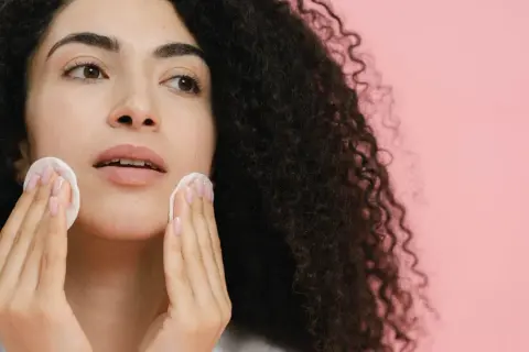 Donna con capelli ricci scuri pulisce il viso con dischetti di cotone, affrontando brufoli e punti neri per una pelle radiosa.