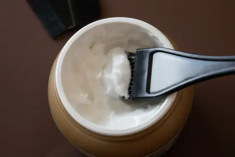 Un pennello raccoglie una crema bianca da un barattolo dorato, ideale per la cura dei capelli con burro di karitè.