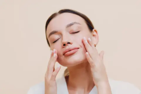 Donna con occhi chiusi applica delicatamente una crema sebo regolatrice sul viso, godendosi un momento di cura della pelle.