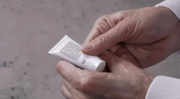 Mani che stringono un tubetto di crema, forse un trattamento iodase anticellulite tiroide, leggendo attentamente gli ingredienti.