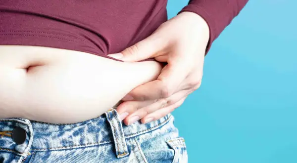 Mano che stringe la pancia, evidenziando gonfiore e possibile ritenzione idrica pancia. Jeans a vita bassa e maglia bordeaux.