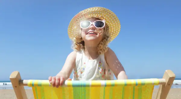 Bambina sorridente con cappello di paglia e occhiali da sole, seduta su una sdraio. Posso usare la crema solare per bambini per proteggerla dal sole.