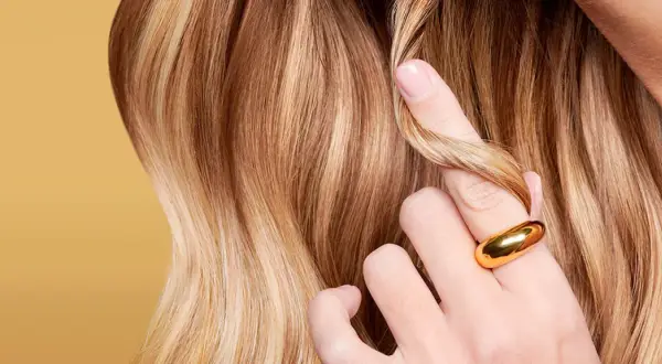 Capelli biondi con riflessi dorati, una mano con anello sfiora una ciocca. Il riflessante copre i capelli bianchi, donando luminosità.