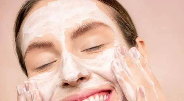 Donna sorridente con maschera bianca sul viso, mentre impara come fare una pulizia del viso a casa.