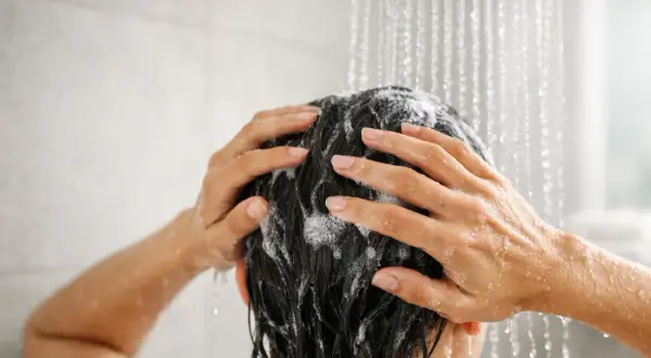 Mani che massaggiano shampoo sui capelli scuri sotto la doccia. Lavare i capelli tutti i giorni fa male, meglio farlo meno spesso.