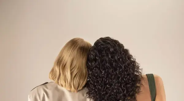 Due donne, una con capelli biondi lisci e l'altra con capelli scuri ricci, si abbracciano. La domanda è quanto dura una tinta permanente.