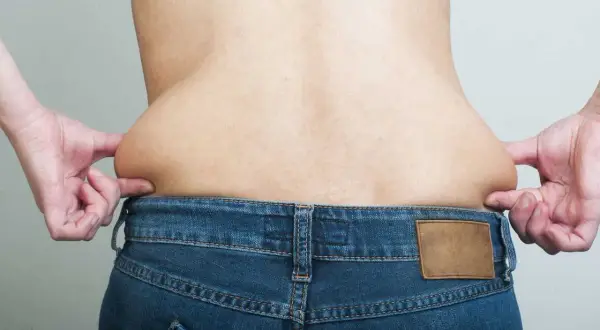 Mani che stringono i fianchi larghi di un uomo, evidenziando accumuli di grasso sopra i jeans.