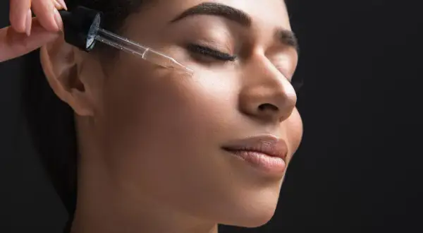 Una mano applica un siero viso con contagocce, forse contenente acido azelaico e retinolo, su una donna con gli occhi chiusi.