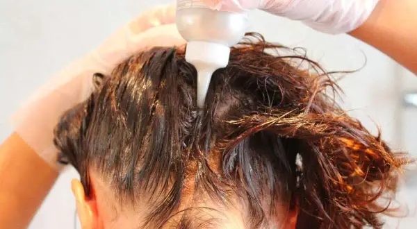 Mani guantate applicano un prodotto per capelli, forse una tinta o un trattamento, direttamente sul cuoio capelluto. Si intravede il flacone con beccuccio.
