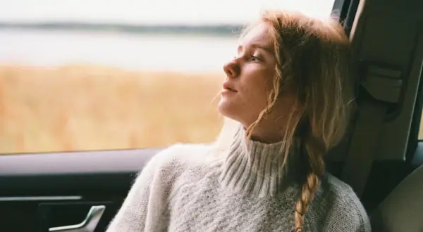 Ragazza con capelli biondi raccolti in acconciature trecce laterali, guarda fuori dal finestrino dell'auto, immersa nei suoi pensieri.