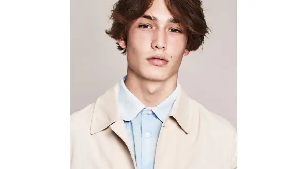 Ragazzo con capelli castani mossi e acconciature alla moda, indossa una camicia azzurra e una giacca beige.
