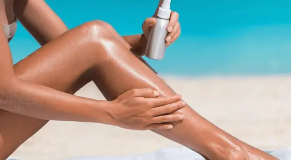 Donna applica spray abbronzante sulle gambe sulla spiaggia, per un'abbronzatura perfetta.