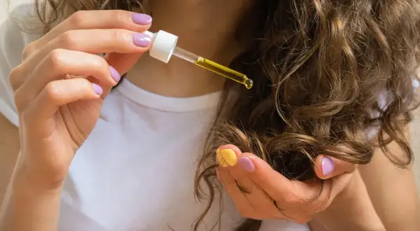 Mani con unghie colorate applicano oli naturali per capelli con un contagocce su ciocche ondulate.