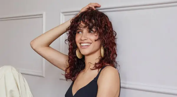 Donna sorridente con capelli castani mossi e tinta rossa, indossa orecchini dorati.