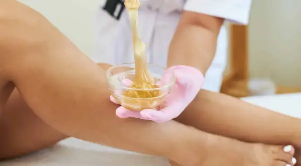 Una mano con guanto rosa preleva cera da una ciotola per la depilazione femminile delle gambe.