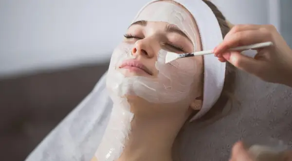 Una donna riceve un peeling viso. La maschera bianca viene applicata con un pennello, mostrando come si fa un trattamento di bellezza.