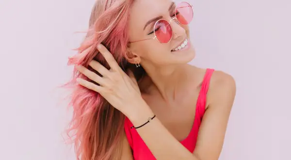 Ragazza con capelli rosa sfumati, occhiali da sole rotondi e un top fucsia. Il suo shampoo colorante mantiene vibrante la chioma.