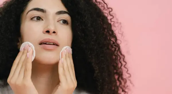 Donna con capelli ricci scuri pulisce il viso con dischetti di cotone, affrontando brufoli e punti neri per una pelle radiosa.