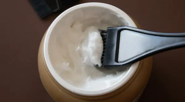 Un pennello raccoglie una crema bianca da un barattolo dorato, ideale per la cura dei capelli con burro di karitè.