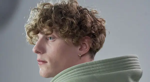 Primo piano di un giovane con capelli mossi uomo, occhi azzurri e un orecchino a cerchio. Indossa una felpa verde chiaro.
