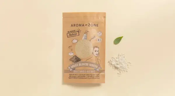 Confezione di maschera argilla bianca Aroma-Zone, con polvere sparsa e fogliolina verde su sfondo chiaro.