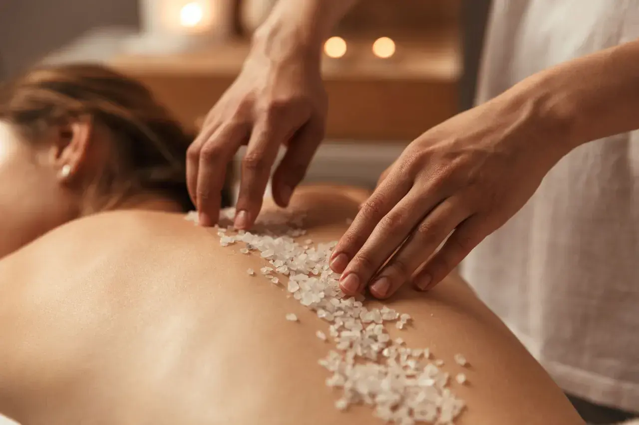 Mani esperte applicano un scrub corpo con cristalli di sale sulla schiena di una persona, un rituale di benessere prima della doccia.