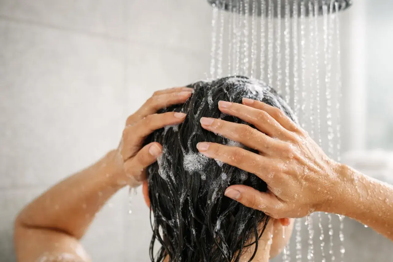 Mani che massaggiano shampoo sui capelli scuri sotto la doccia. Lavare i capelli tutti i giorni fa male, meglio farlo meno spesso.