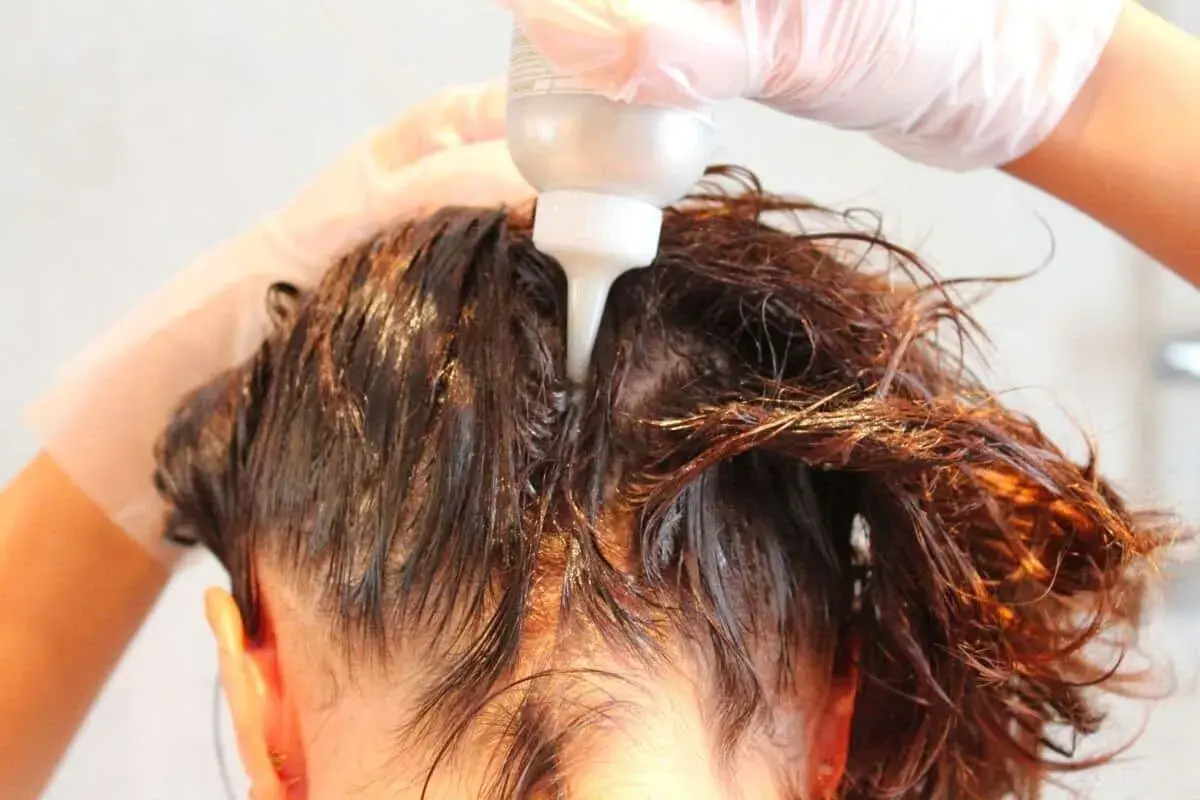 Mani guantate applicano un prodotto per capelli, forse una tinta o un trattamento, direttamente sul cuoio capelluto. Si intravede il flacone con beccuccio.
