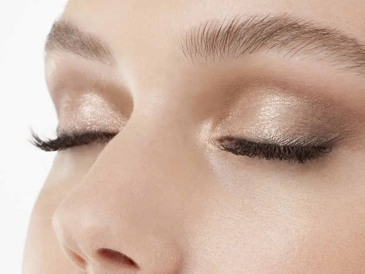 Occhi chiusi con trucco luminoso, ciglia lunghe e folte. Uno sguardo spento che riflette un momento di quiete interiore.