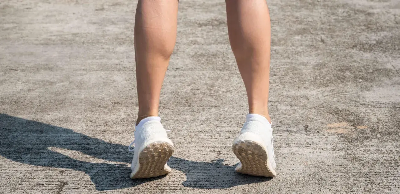 Gambe di donna con polpacci grossi, forse a causa di allenamento intenso. Scarpe da ginnastica bianche su cemento.