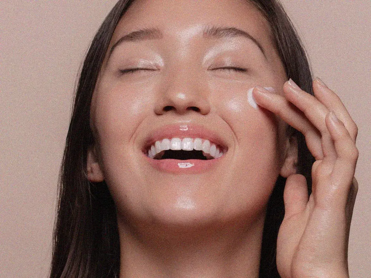 Viso radioso di una donna che applica una crema, un gesto che ricorda cos'è lo scrub per una pelle luminosa e liscia.